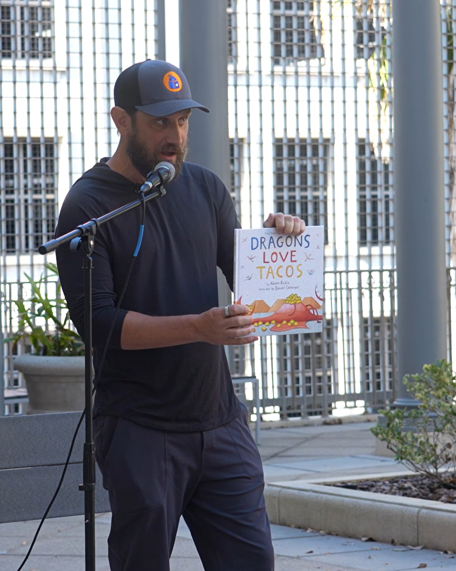 Rich reading Dragons Love Tacos outdoors at a mic
